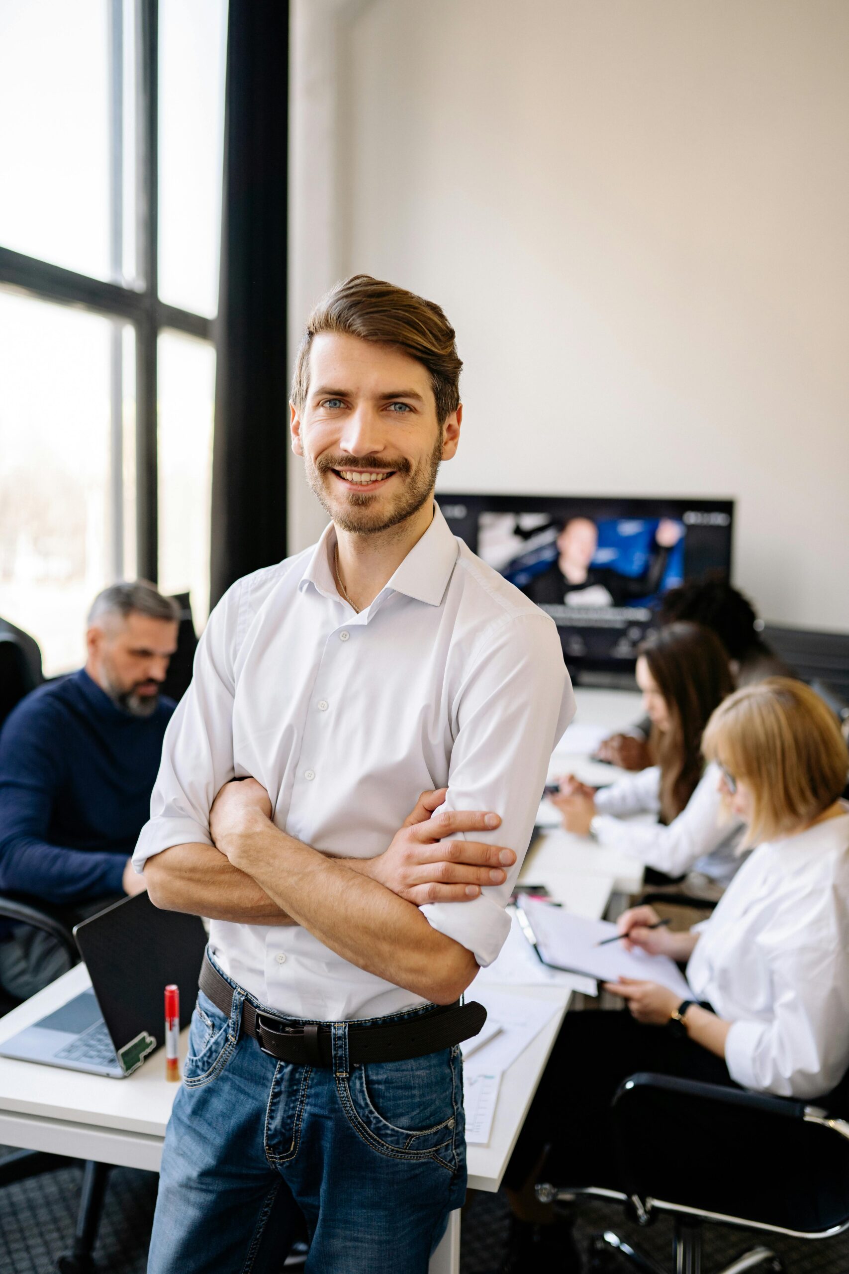 Smiling man standing confidently in a modern office environment with colleagues working in the background.